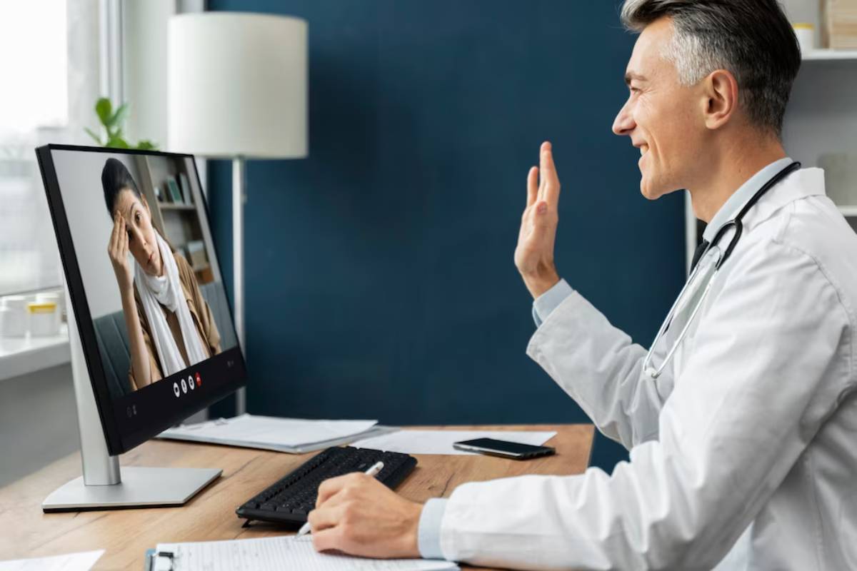 A doctor gestures while conducting a virtual consultation with a patient on a computer screen in a well-lit office.