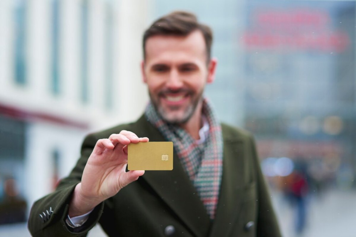 A person holds a shiny gold card in front of them, dressed in a dark coat and a patterned scarf, with a blurred urban background.
