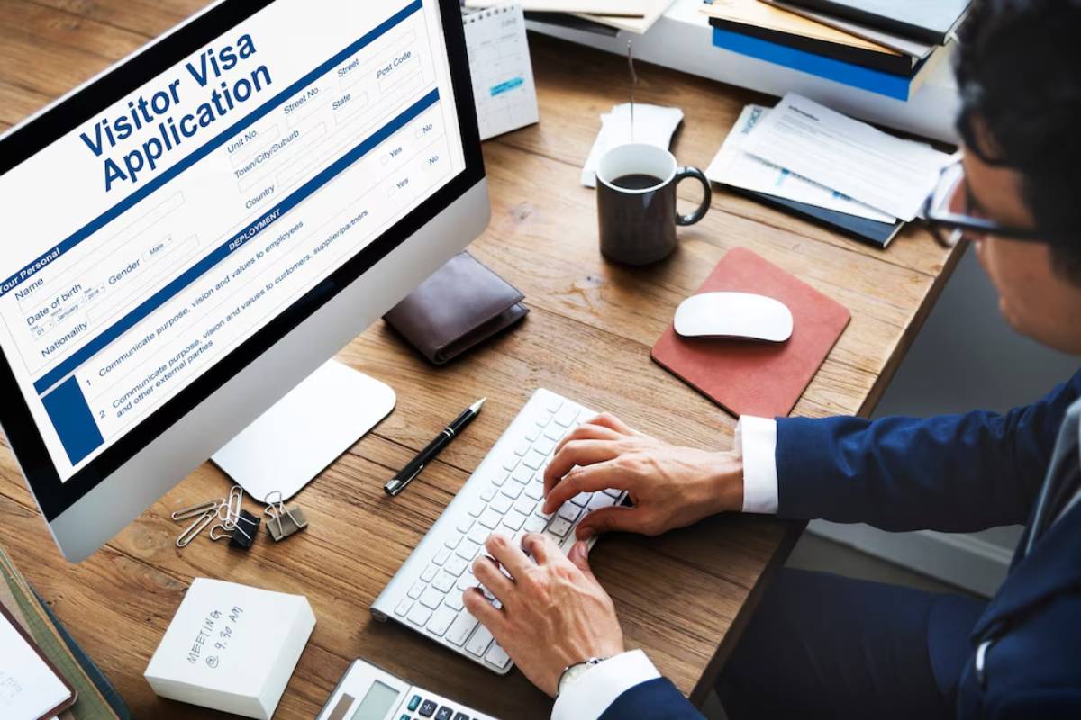 A person in a suit filling out a Visitor Visa application on a computer, surrounded by paperwork and a coffee mug on a wooden desk.