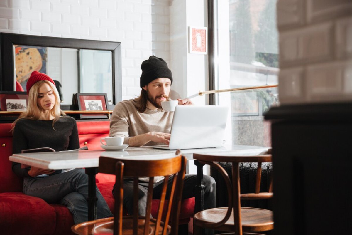 Two people sit in a cozy café, one working on a laptop while the other reads, surrounded by simple wooden furniture and a bright atmosphere.