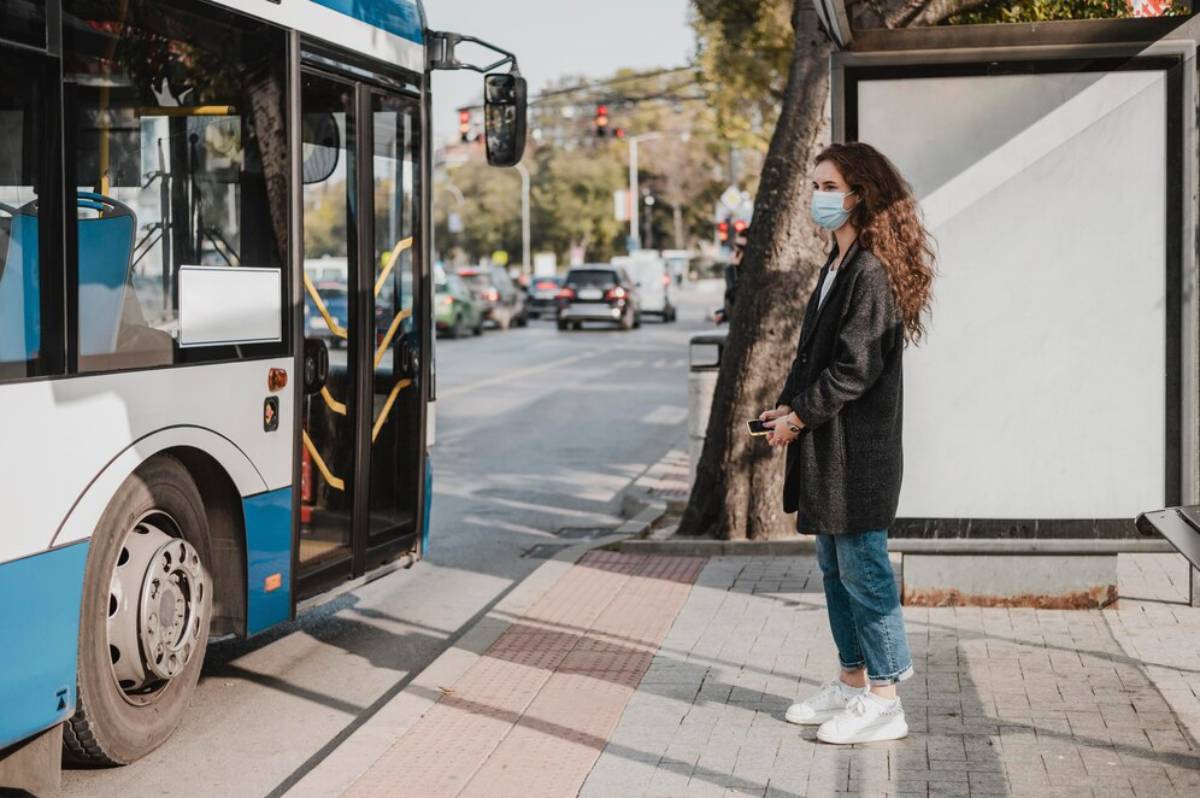 A woman stands at a bus stop, dressed in a long black coat and holding a phone, while a blue and white bus approaches.
