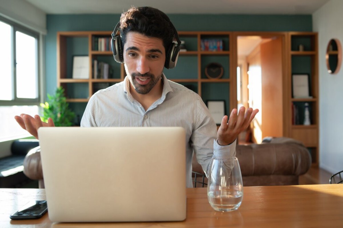 A person in a striped shirt wearing headphones sits at a desk, gesturing towards a laptop, with a glass of water beside them.