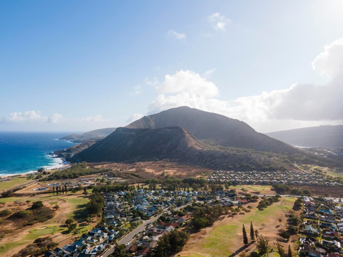 Aerial view of rugged coastline with a mountain backdrop, overlooking a residential area and golf course beneath a bright blue sky.