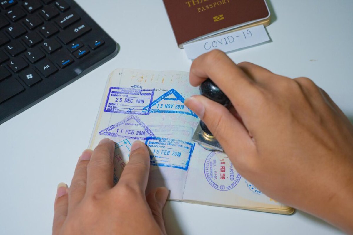 A hand holding a stamp presses down on a passport with multiple entry stamps, beside a keyboard and a labeled COVID-19 note.