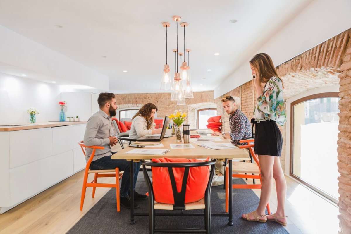 A modern meeting space with four people gathered around a wooden table, engaged in discussion amidst bright decor and soft seating.