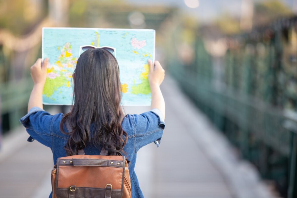 A traveler with long hair holds a map while standing on a bridge, wearing a denim jacket and a brown backpack.