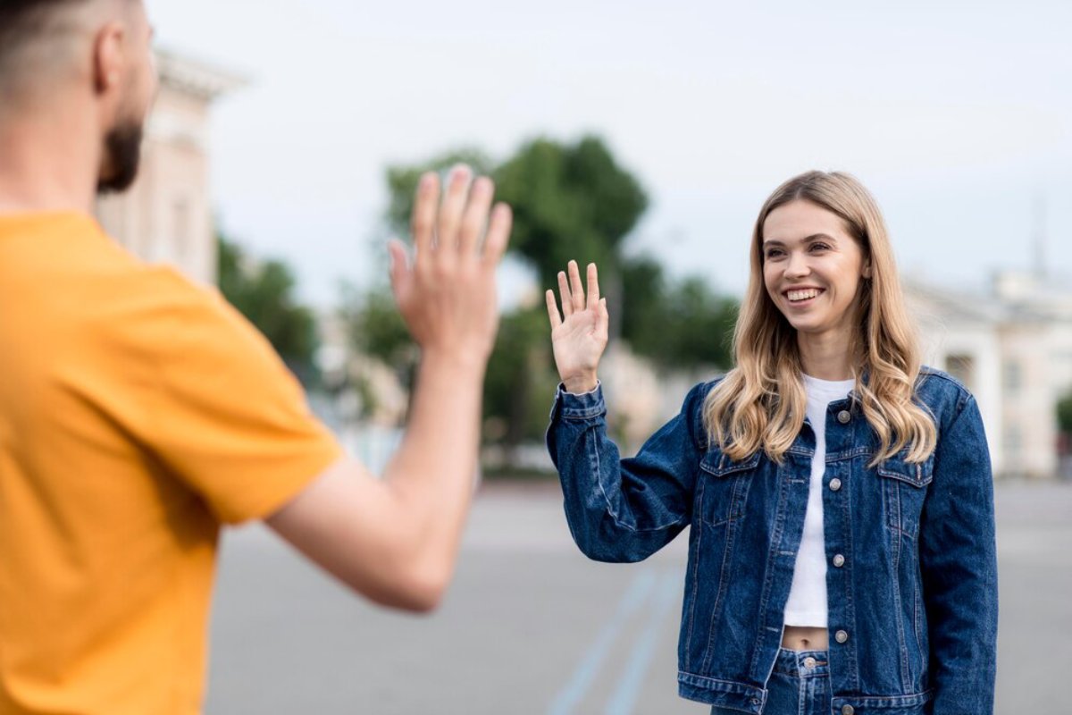 A man in an orange shirt and a woman in a denim jacket wave at each other in an outdoor setting.