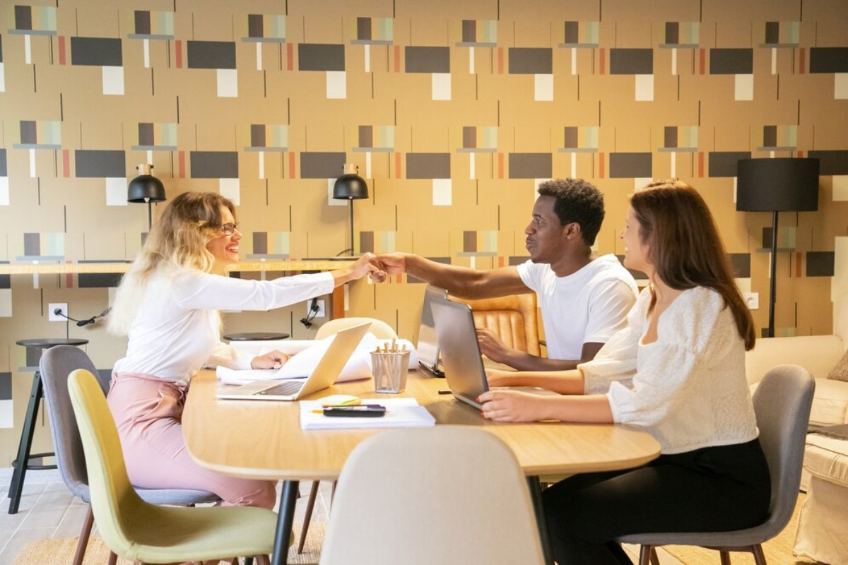 A lively business meeting with three professionals engaging at a round table, laptops and documents present, set against a stylish background.