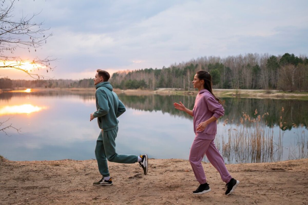 A man and woman jog along a sandy lake shore at sunset, surrounded by trees and a peaceful, reflective water surface.