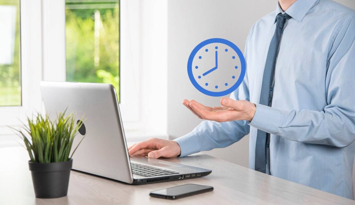 A businessman in a blue shirt holds his hand out, displaying a blue clock icon, with a laptop and plant on his desk.