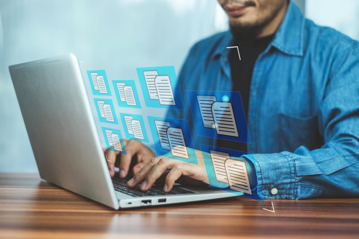 A man in a denim shirt types on a laptop, surrounded by floating icons of documents, symbolizing digital document management.