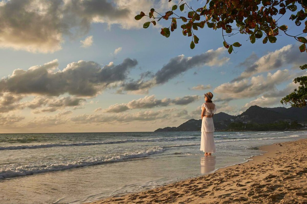 A woman in a white outfit stands on a beach, gazing at the ocean under a cloudy sky during sunset.