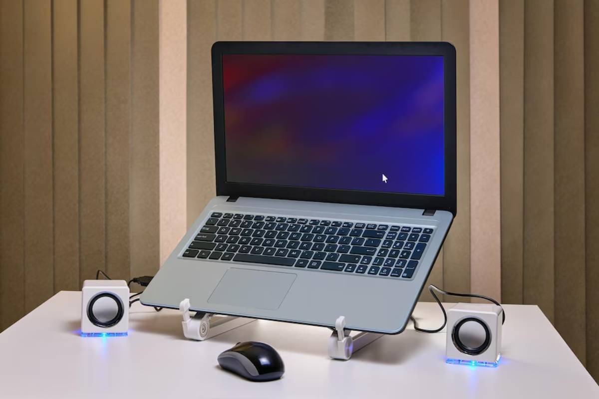 A laptop with a blank screen sits on a stand, flanked by two small white speakers and a black mouse, against a striped backdrop.