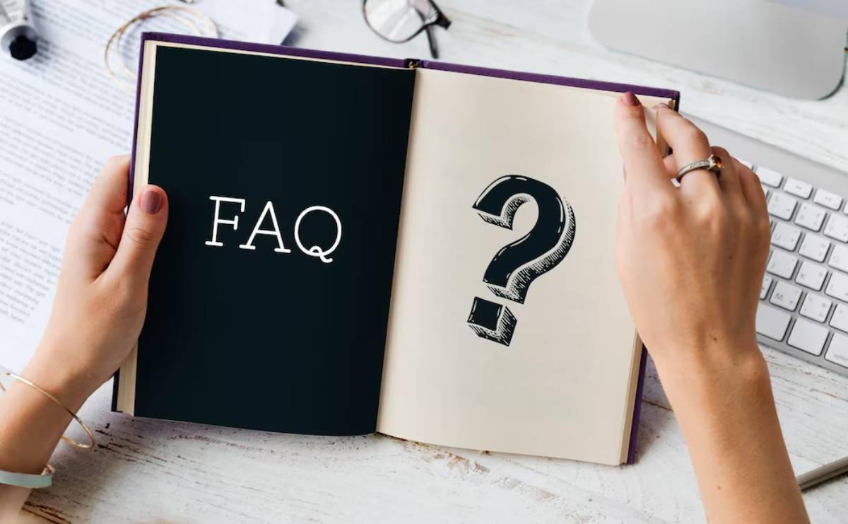 A hand holds an open book displaying FAQ and a large question mark, against a backdrop of office supplies.