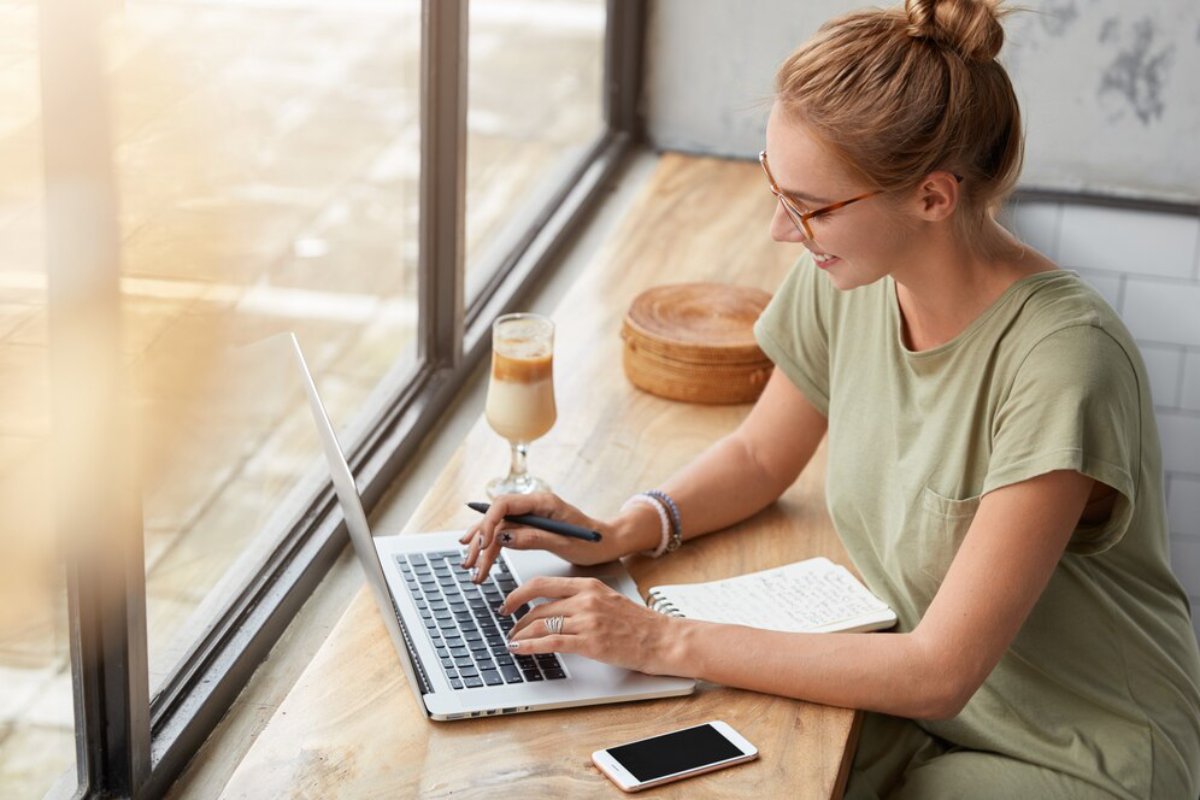 A woman in a green t-shirt works on a laptop at a wooden table, accompanied by a notebook and a glass of iced coffee.