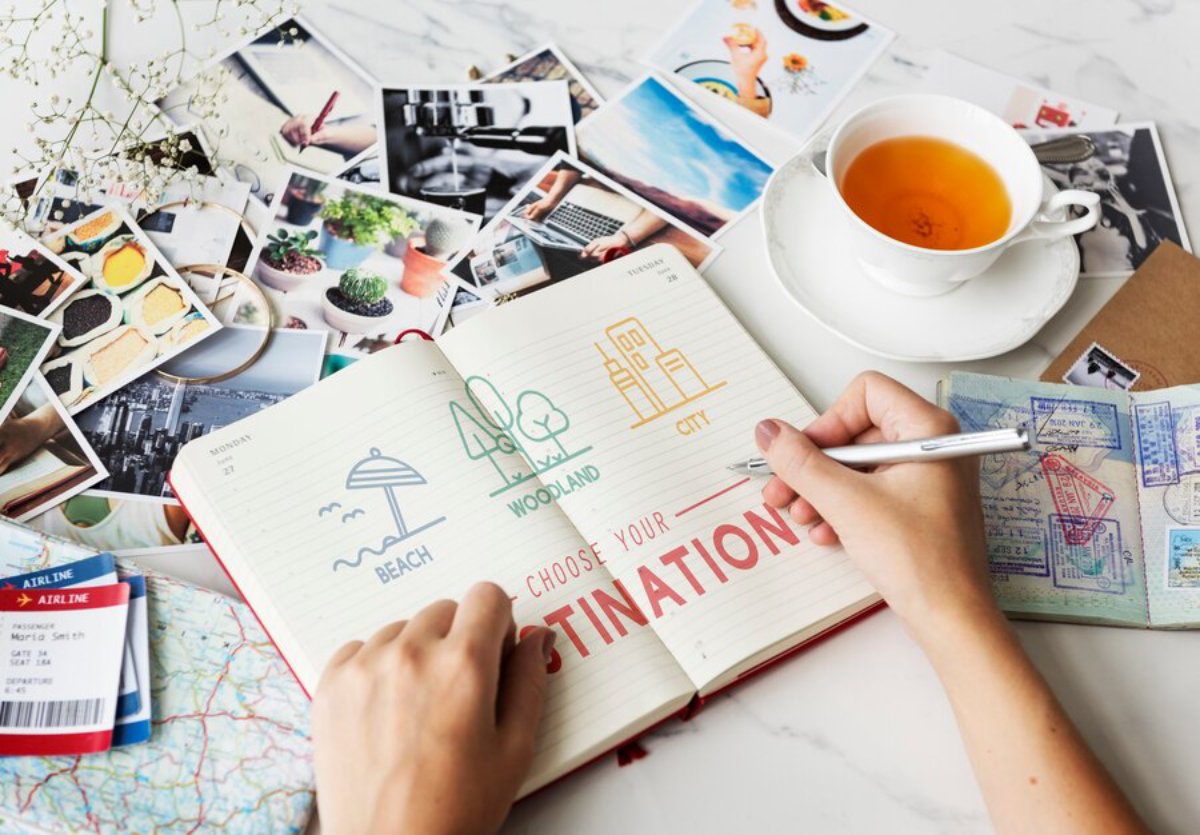 A hand holding a pen writes Choose Your Destination in a planner, surrounded by travel photos, a cup of tea, and a map.
