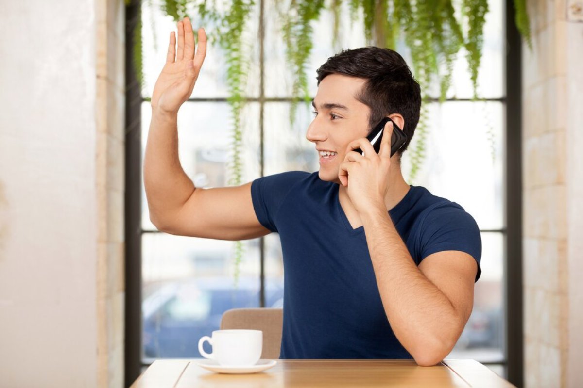 A person with a smartphone at a café table raises a hand while engaged in a conversation, with plants visible in the background.