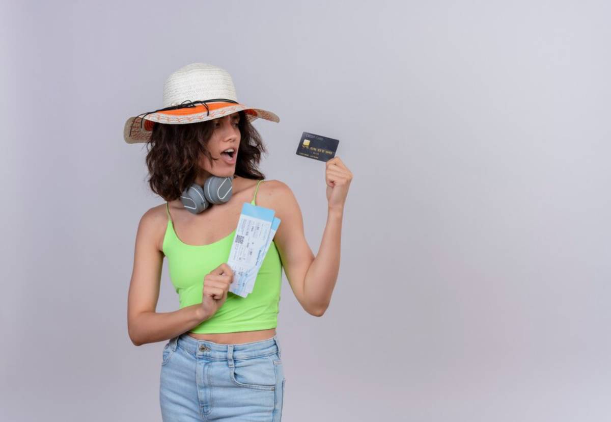 A person in a green tank top holds a boarding pass and a credit card, wearing headphones and a sun hat, against a plain background.