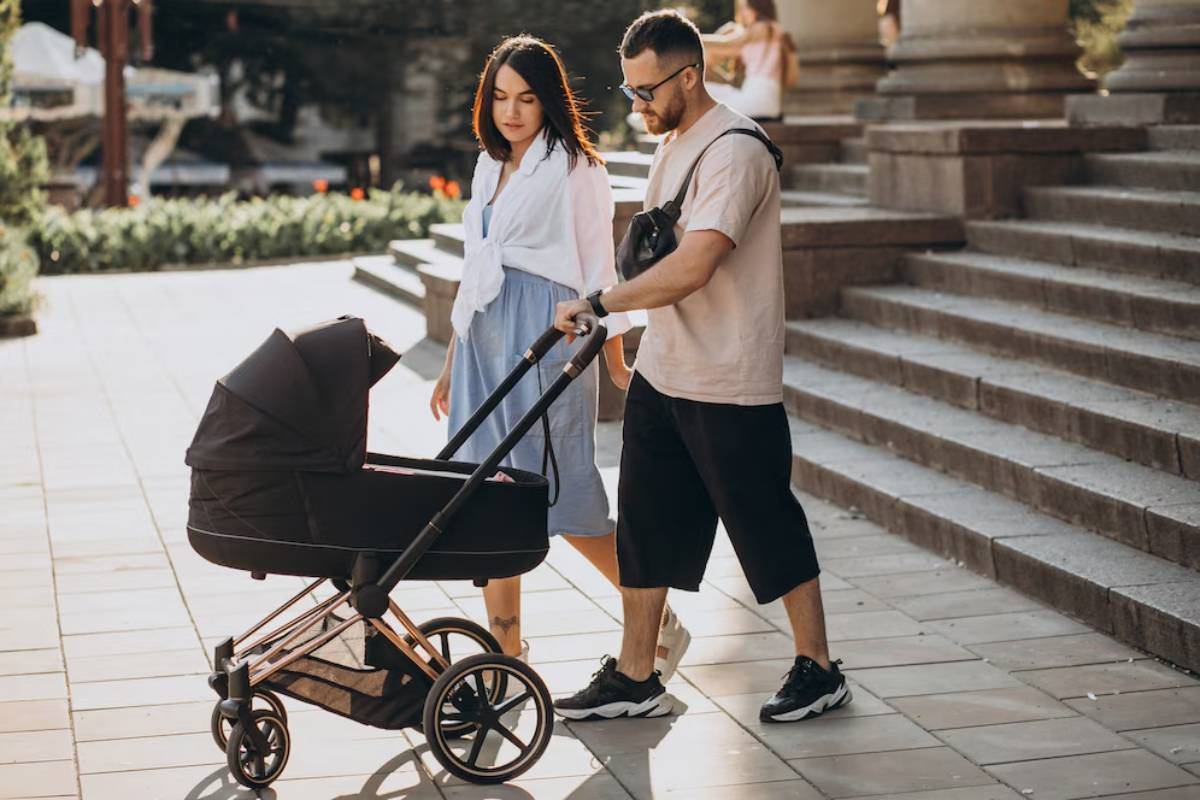 A couple strolls together, one pushing a sleek black stroller along a stone pathway, with greenery and steps in the background.