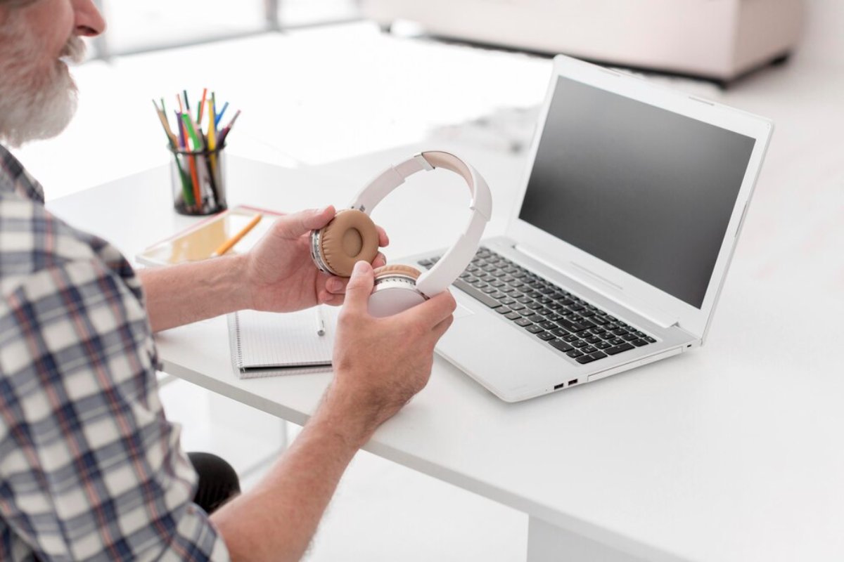 A person holds a pair of white headphones, sitting at a desk with a laptop, notepad, and colorful pens in the background.