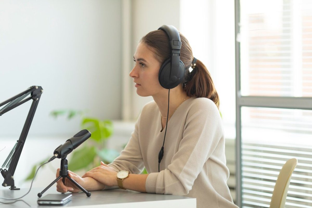 A person wearing headphones sits at a desk with a microphone and has their hands resting on the table, surrounded by natural light.