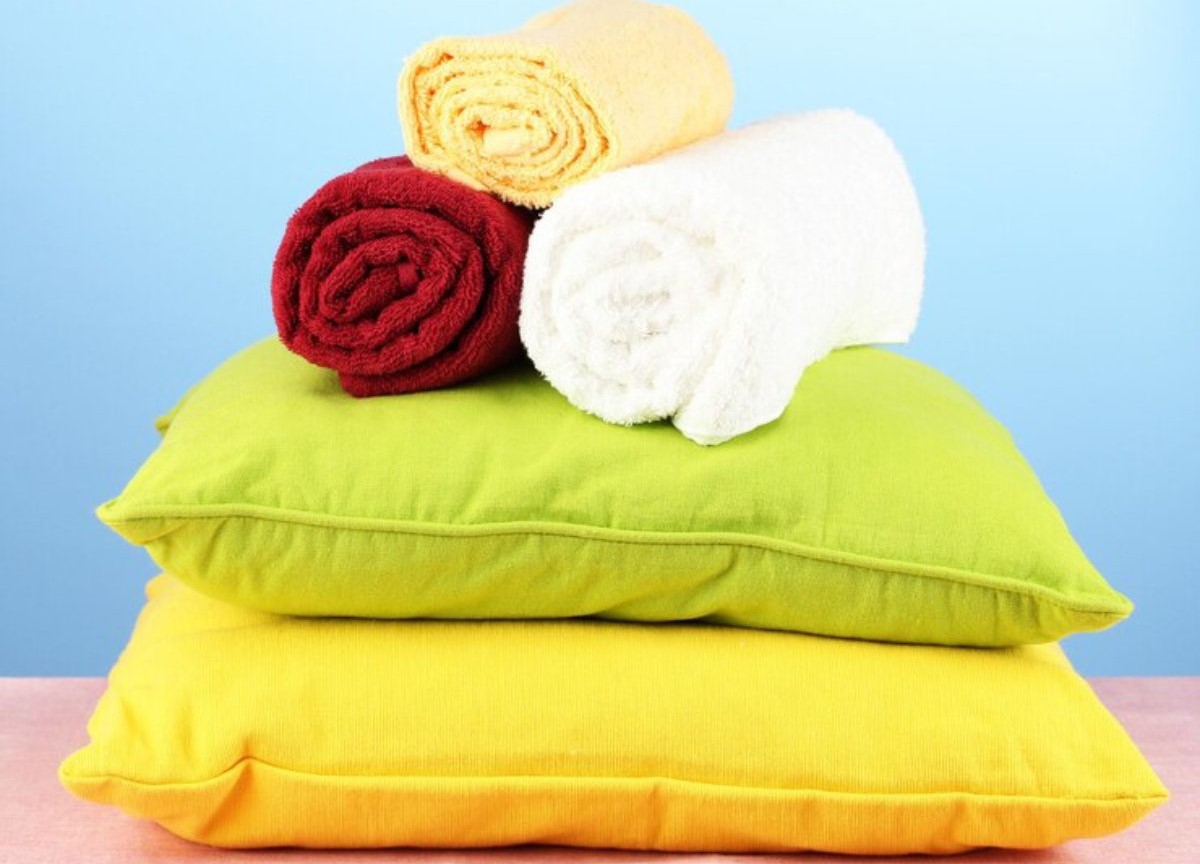 A stack of colorful towels in red, white, yellow, and green rests on two bright cushions, set against a soft blue background.