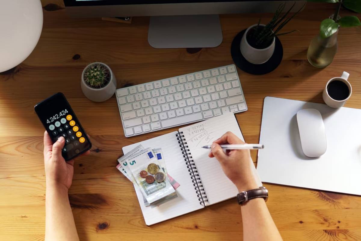 A hand holding a smartphone with a calculator display, writing notes in a notebook, surrounded by cash, a computer, and a coffee cup.