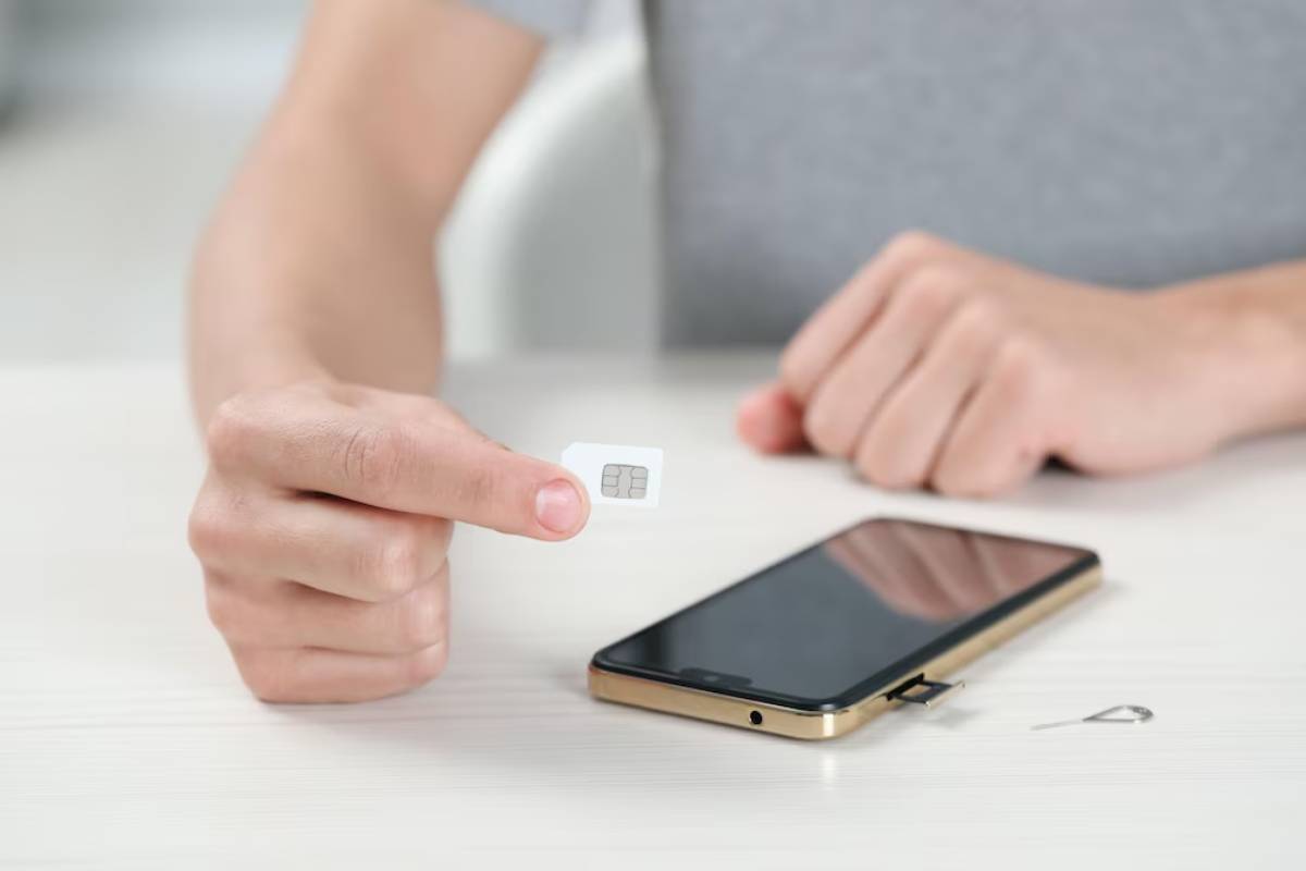 A person holding a SIM card beside a smartphone on a table, with a SIM eject tool nearby.