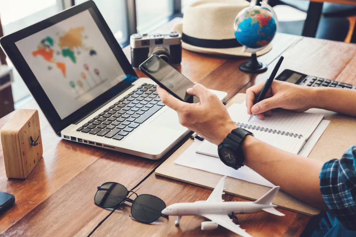 A person uses a smartphone while writing in a notebook, surrounded by a laptop, globe, camera, and travel-themed items.