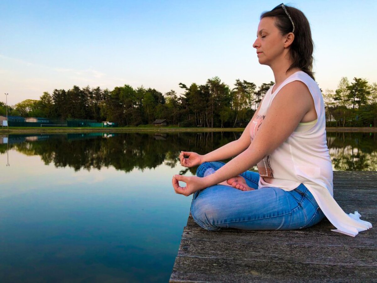 A person sits cross-legged on a wooden dock by a tranquil lake, practicing meditation while surrounded by lush greenery and a clear sky.