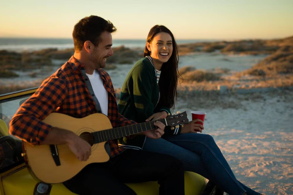 A person strums a guitar while sitting on a yellow car, with a sunset beach scene and a companion holding a red cup beside them.