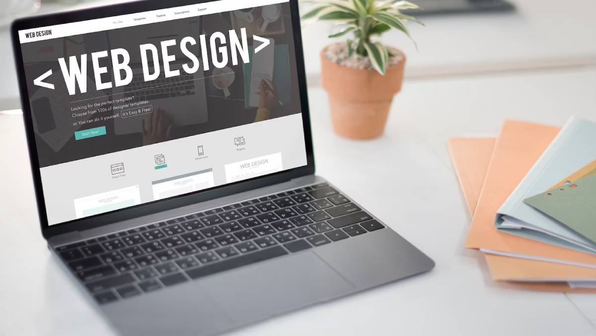 A laptop displays a web design website with a clean layout, surrounded by a potted plant and colorful folders on a desk.
