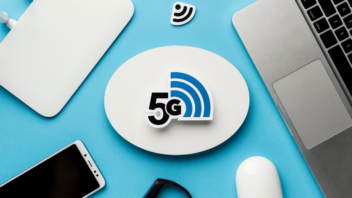 A 5G logo prominently displayed on a white oval, surrounded by a laptop, smartphone, mouse, and other tech accessories on a blue background.