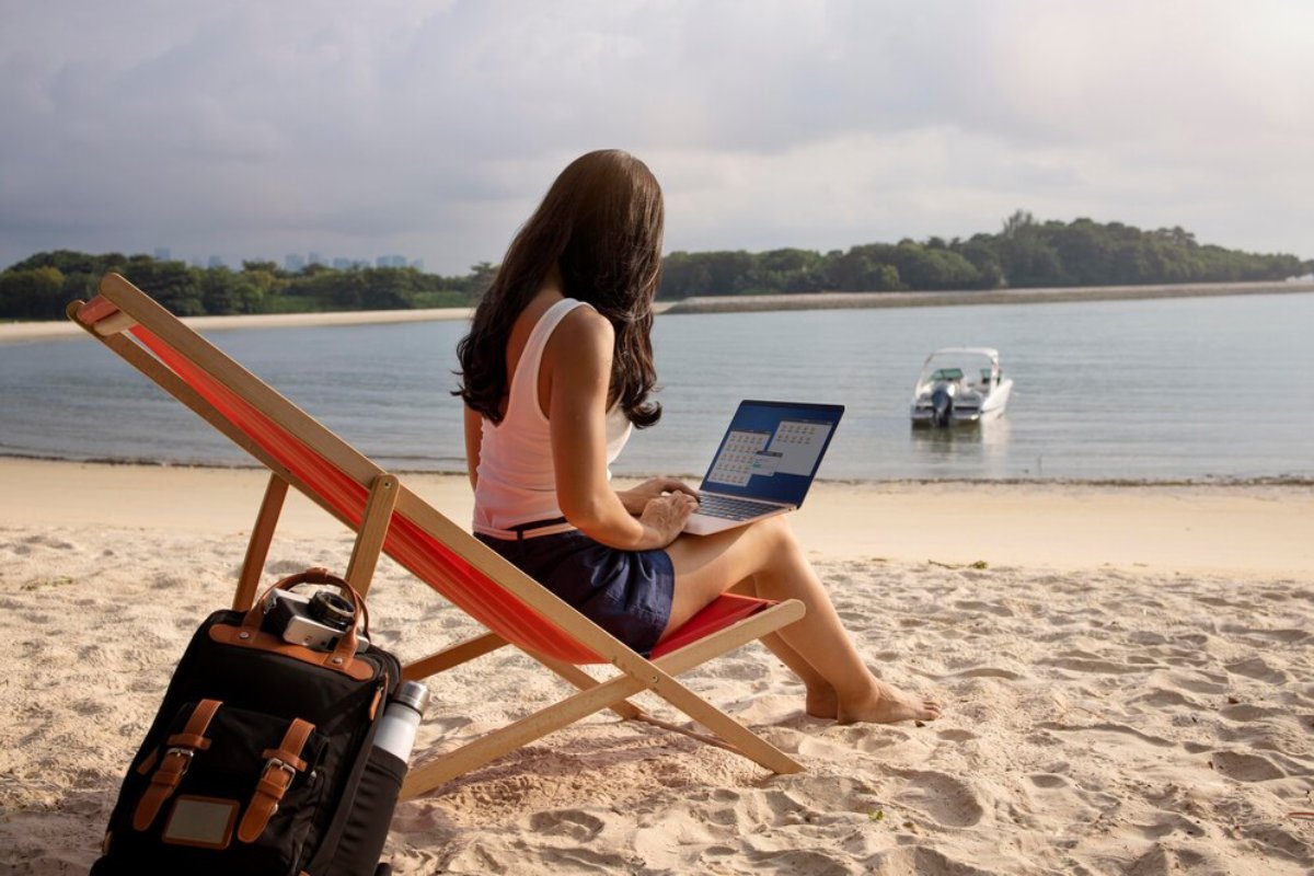 A woman sitting on a beach chair works on her laptop, with a serene ocean view and a boat in the background. A backpack rests nearby.