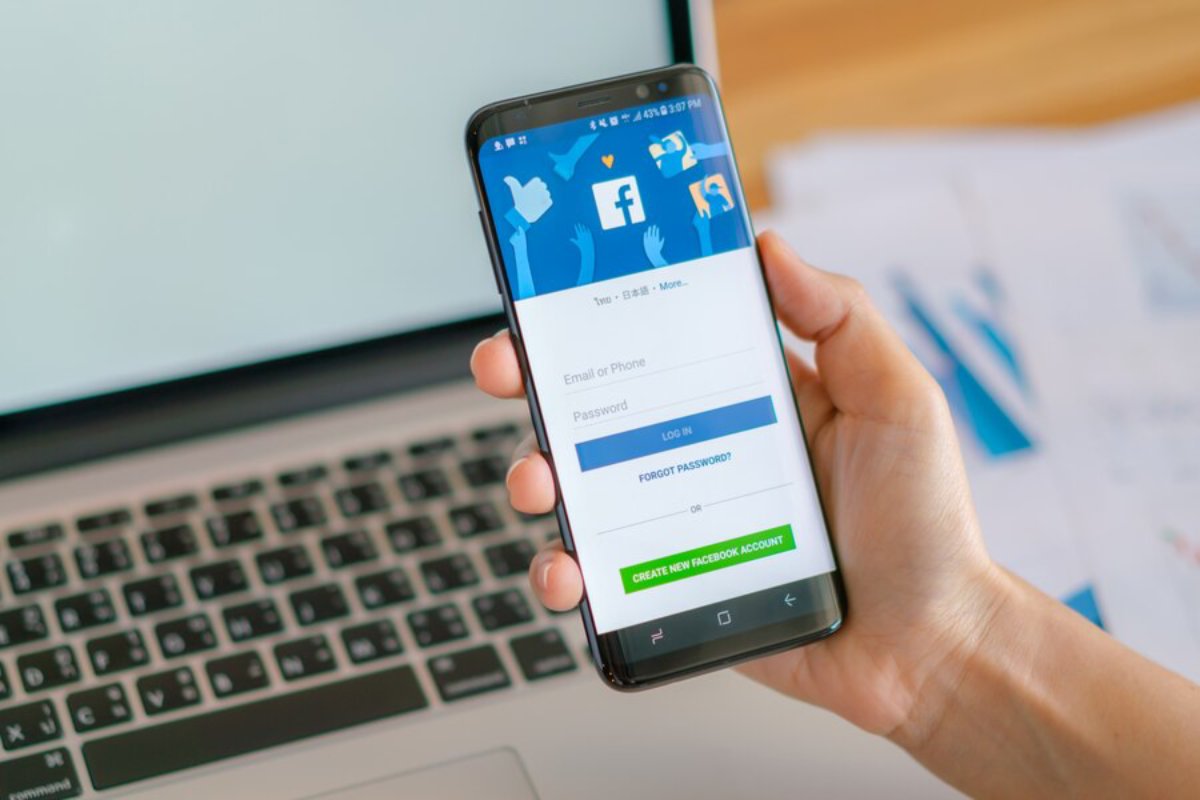 A person holds a smartphone displaying the Facebook login screen, with options for email/phone and password entry.