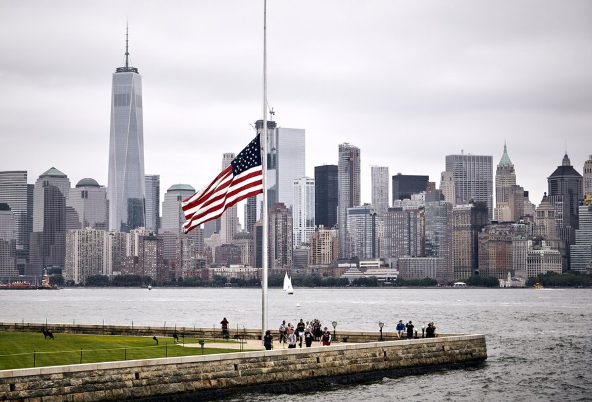 A cloudy New York skyline with One World Trade Center, an American flag at half-mast, and visitors on a waterfront promenade.