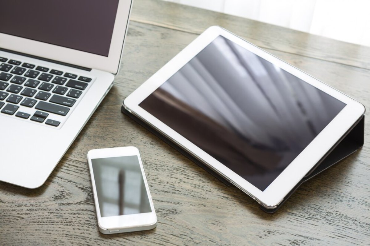A laptop, tablet, and smartphone with blank screens on a wooden desk, illuminated by natural light from a nearby window.