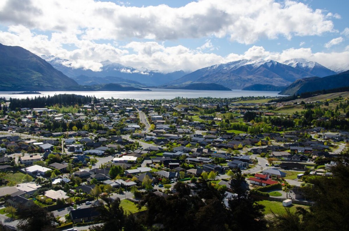 Aerial view of a scenic lakeside town surrounded by mountains, featuring lush greenery, homes, and winding roads under a cloudy sky.