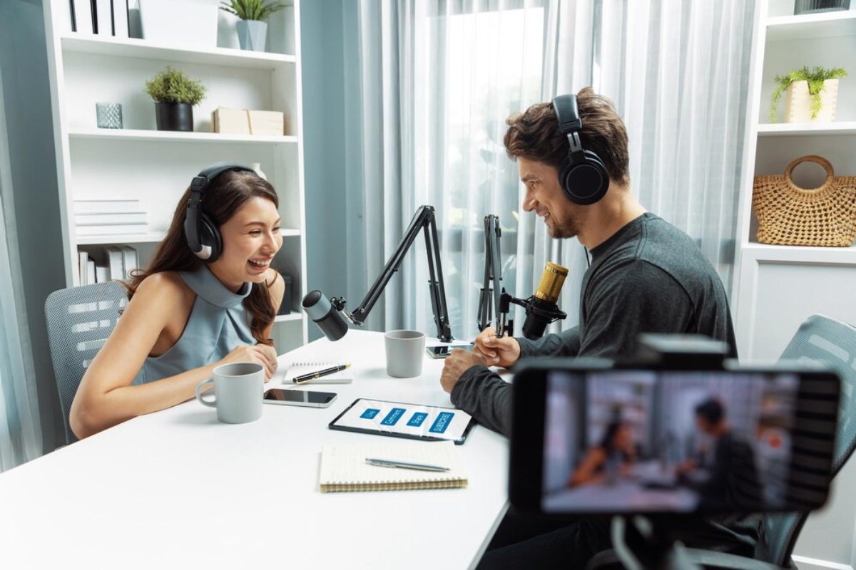 Two individuals are seated at a table with microphones and headphones, engaged in a podcast recording within a modern office space.