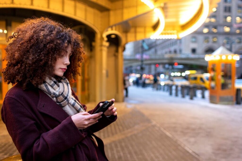 A woman with curly hair checks her phone while standing on a bustling city street, illuminated by warm lights and urban scenery.