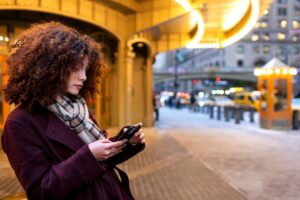A woman with curly hair checks her phone while standing on a bustling city street, illuminated by warm lights and urban scenery.
