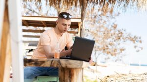 A person seated at a rustic table working on a laptop beside a beach, with a drink beside them, under a straw umbrella.