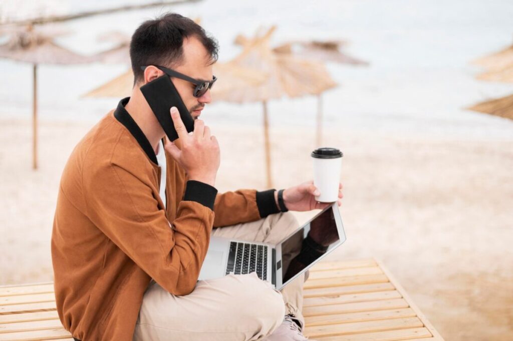 A person sits on a wooden bench by the beach, talking on the phone while holding a coffee cup and working on a laptop.