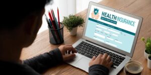 A person fills out a health insurance application on a laptop, surrounded by stationery and a plant on a wooden desk.