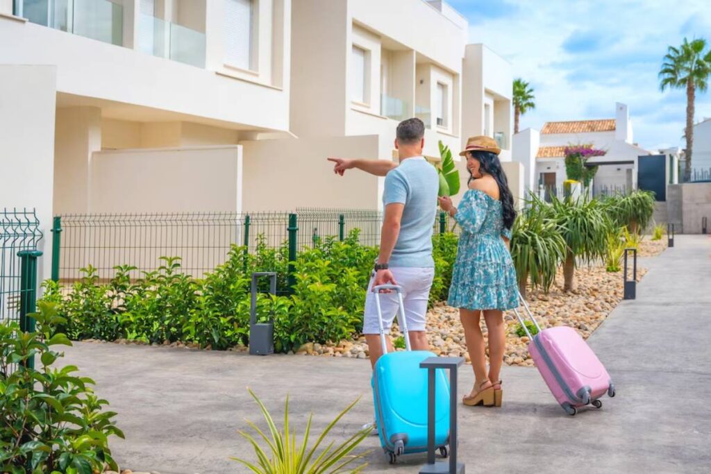 A couple stands in front of modern buildings, pointing towards a destination with colorful luggage in tow amid lush greenery.