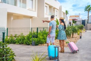 A couple stands in front of modern buildings, pointing towards a destination with colorful luggage in tow amid lush greenery.