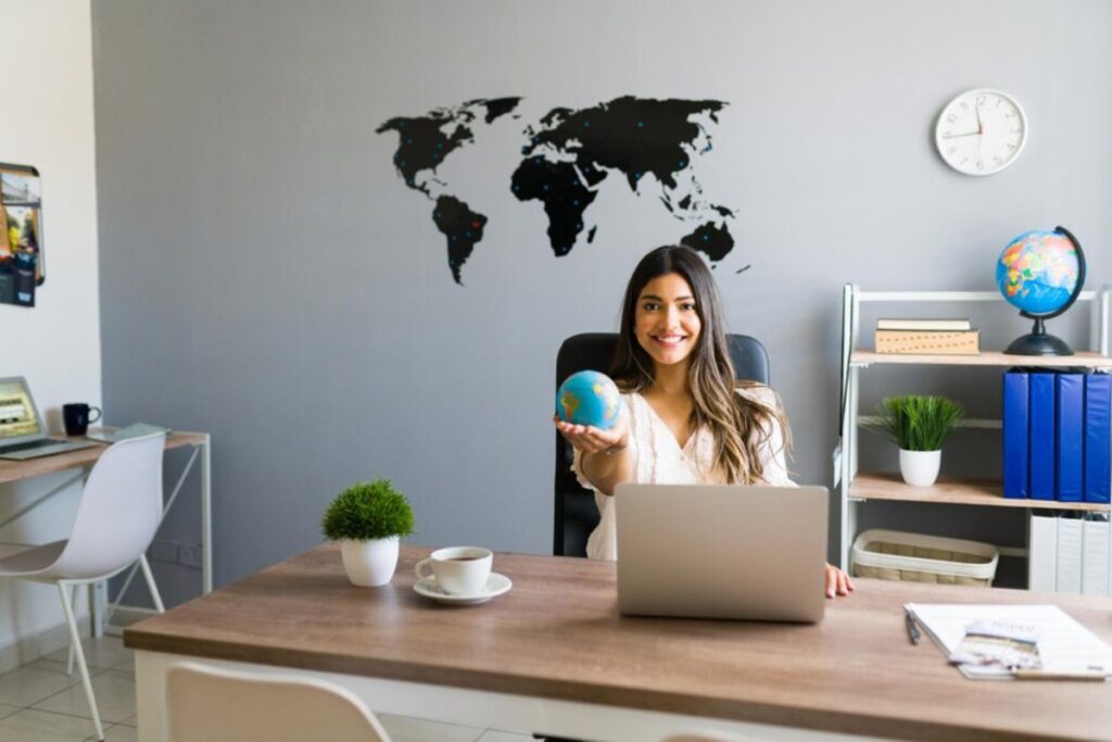 A woman sits at a desk holding a globe, surrounded by office supplies and a world map on the wall, conveying a professional atmosphere.