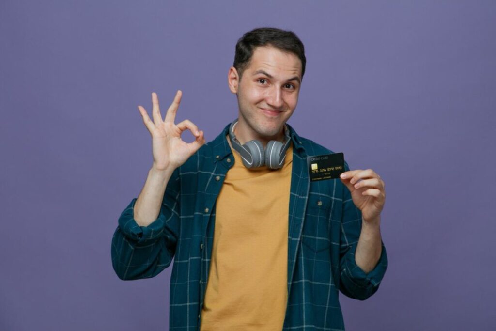 A person wearing headphones and a plaid shirt holds a credit card while making an OK hand gesture against a purple backdrop.