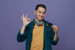 A person wearing headphones and a plaid shirt holds a credit card while making an OK hand gesture against a purple backdrop.