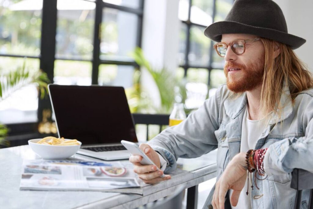 A person with long hair and a hat sits at a table, using a smartphone, with a laptop and a bowl of fries nearby.